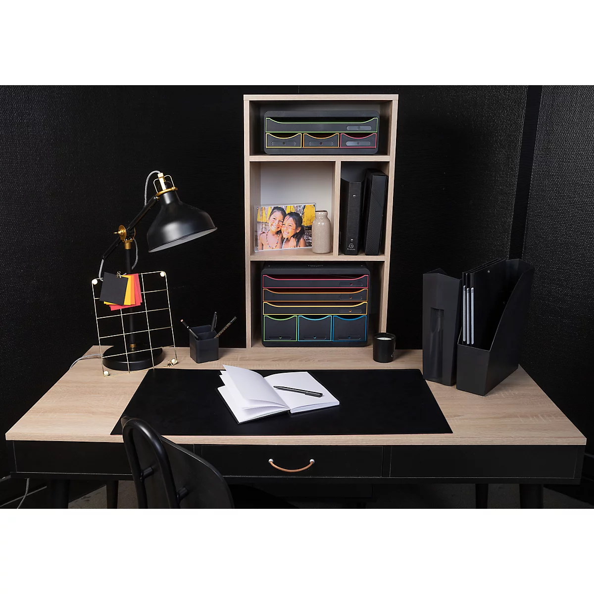 Schreibtisch mit schwarzer Tischplatte, Lampe, Notizblock, Ablagen, Bücher und Accessoires. Schwarzer Hintergrund.