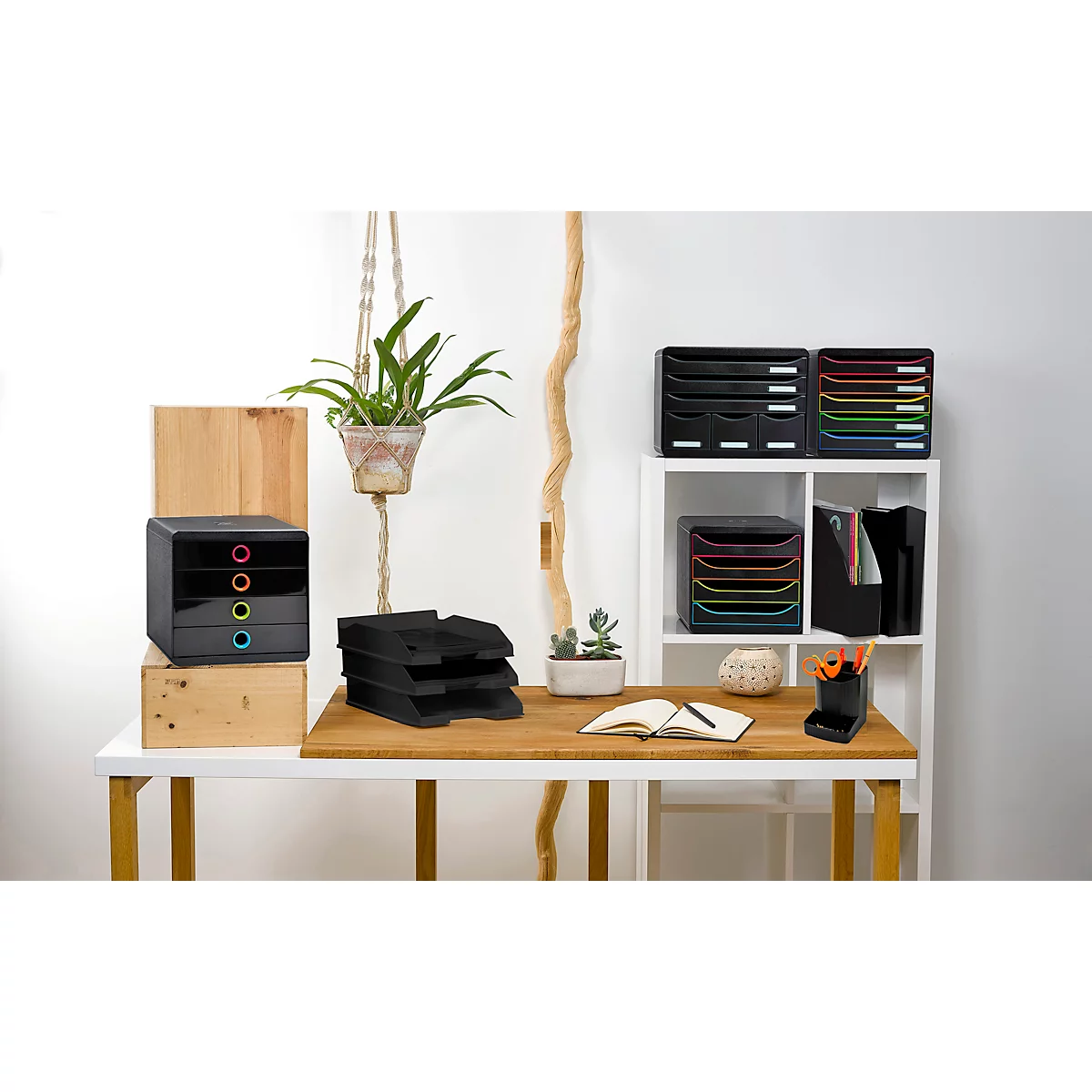 Ein Schreibtisch mit Büromaterialien: schwarze Ablagesysteme, Pflanze, Notizbuch. Verschiedene Farben akzentuieren das Set.