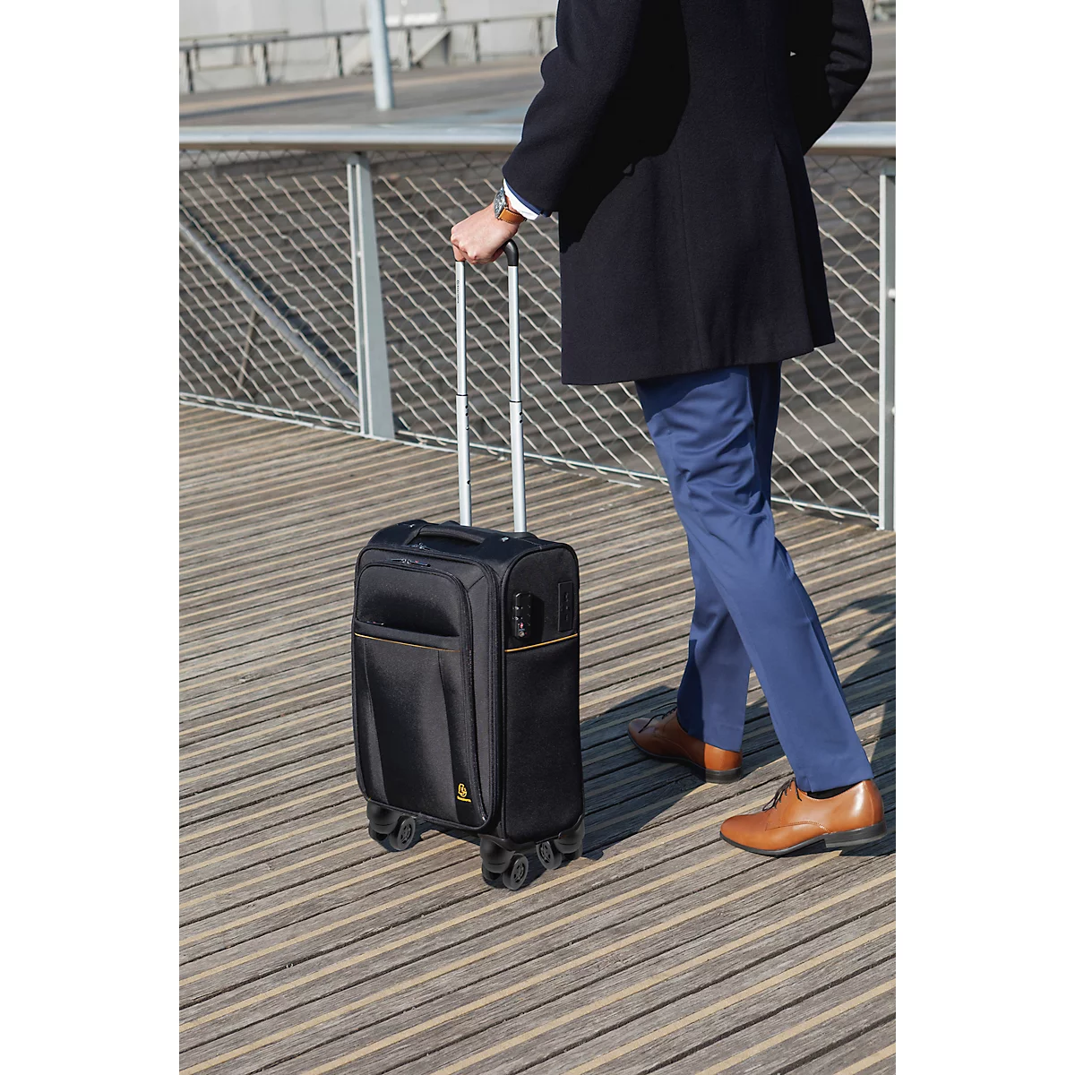 Person zieht einen schwarzen Trolley auf Holzstegen. Er trägt einen dunklen Mantel, blaue Hose und braune Schuhe.