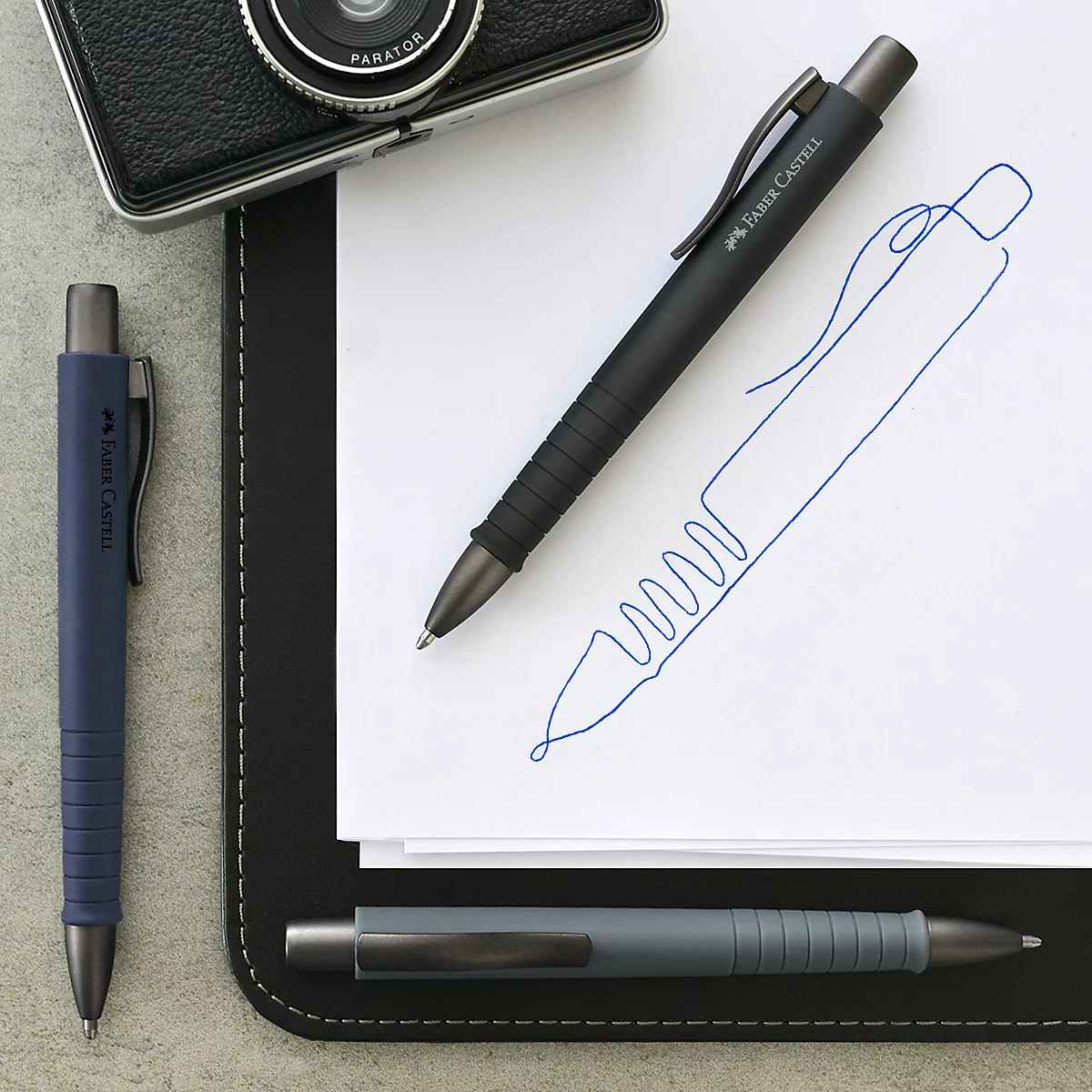 Drie balpennen op een tafel, waaronder een zwarte, een blauwe en een grijze pen.