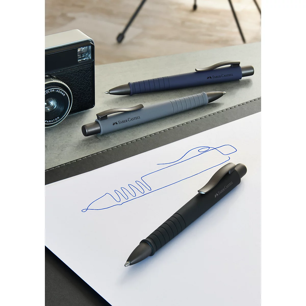 Three pens in blue, grey, and black lie on a surface, with a sketch below.