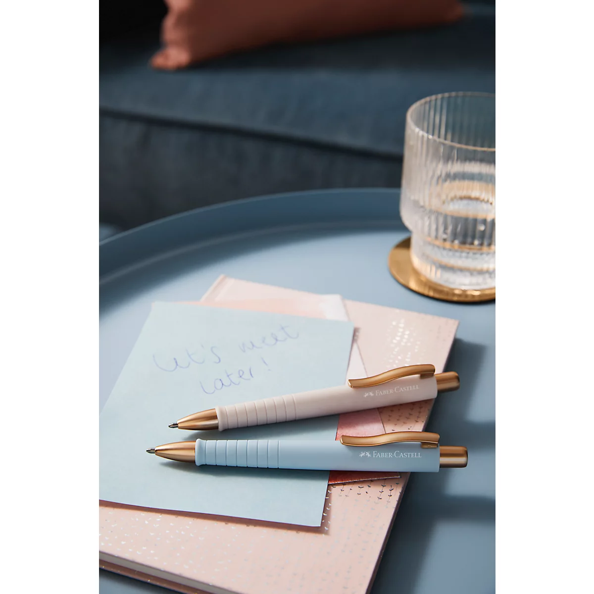 Two pens rest on notepads. A light blue note says: Let's meet later! Next to it is a glass of water.