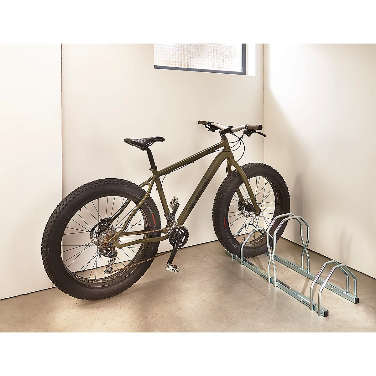 Ein olivgrünes Fatbike steht neben einem Fahrradständer in einer hellen Ecke.