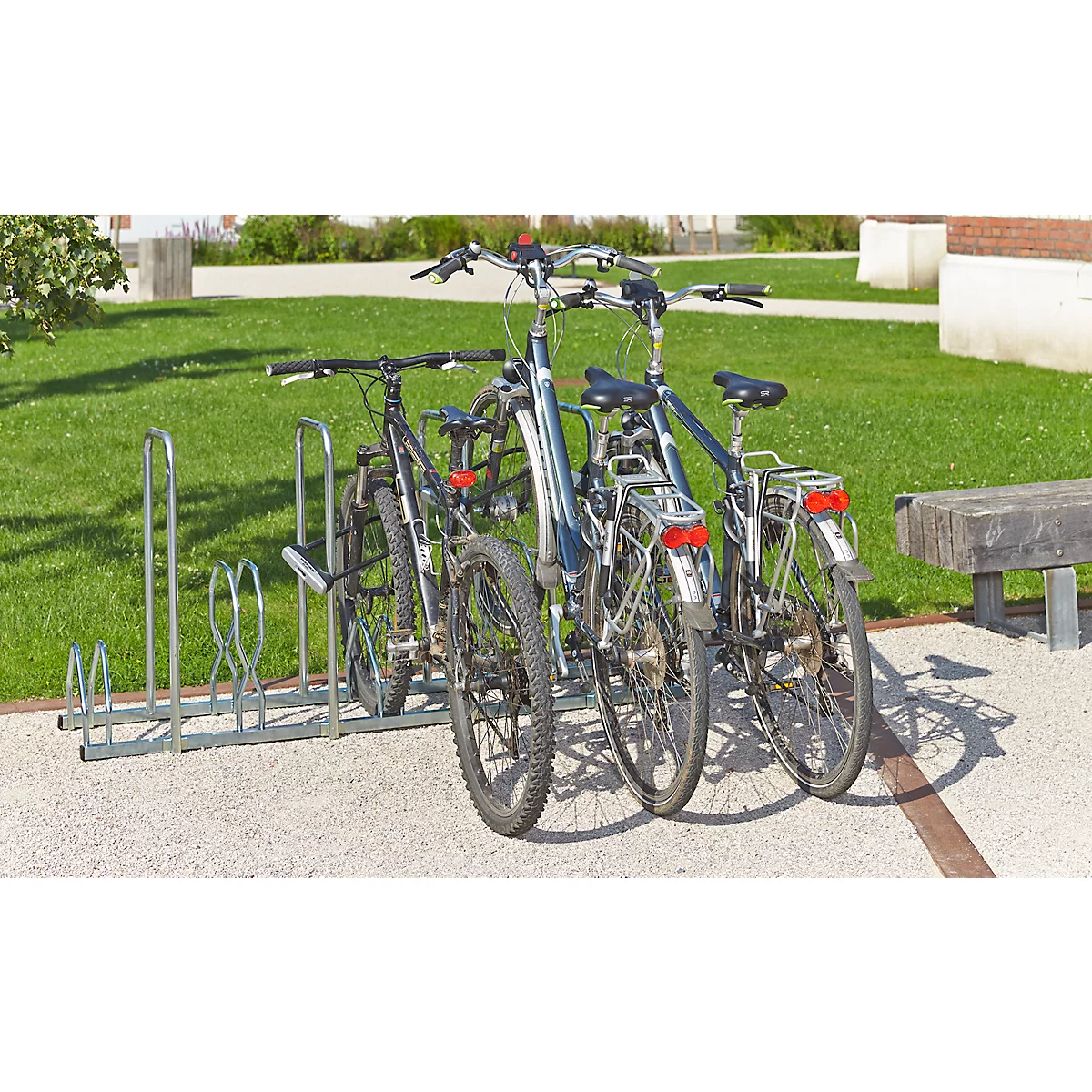 Drei Fahrräder in einem Fahrradständer auf einem gepflasterten Weg. Im Hintergrund Rasen und ein Gebäude.