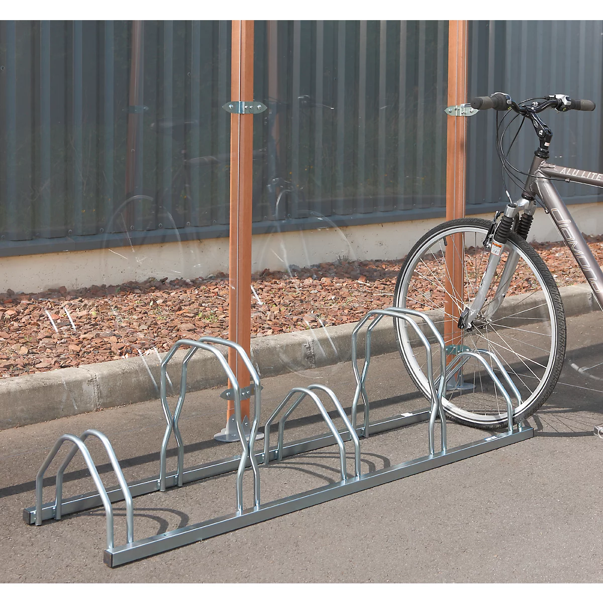 Fahrradständer aus Metall mit Fahrrad davor. Im Hintergrund eine transparente Wand und eine graue Fassade.