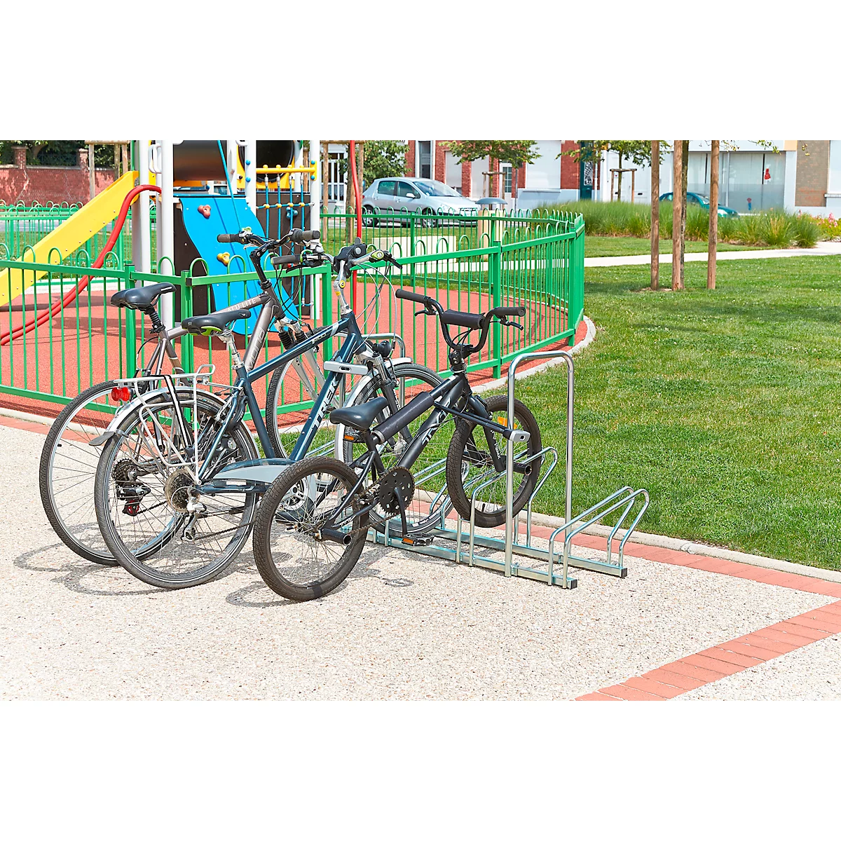 Drei Fahrräder stehen auf einem Gehweg in einem Fahrradständer. Im Hintergrund befindet sich ein Spielplatz.
