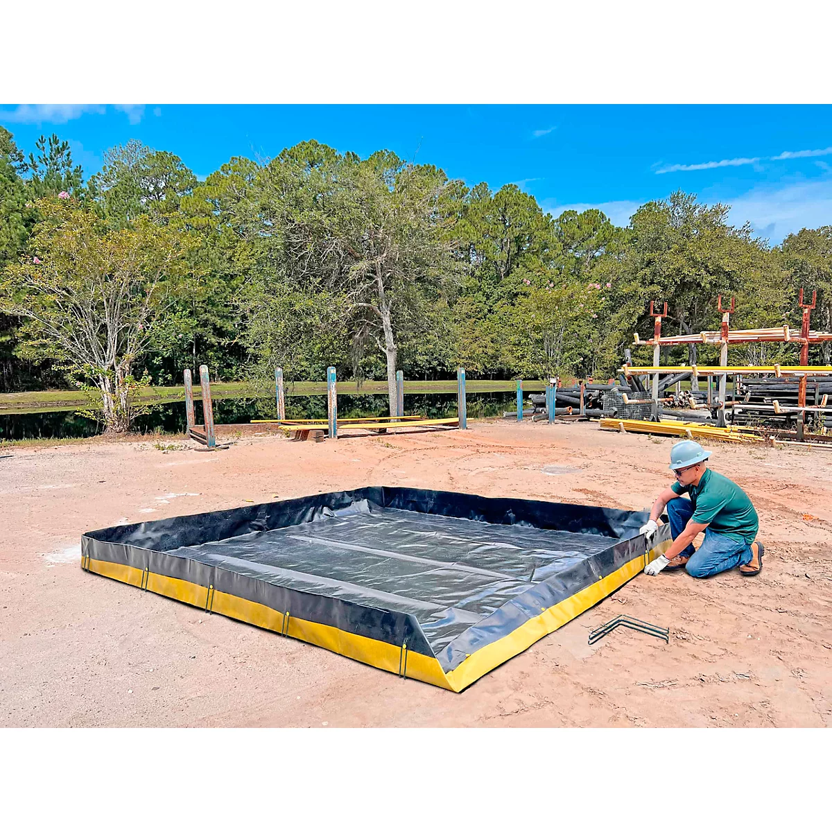 Ein Arbeiter installiert eine schwarze und gelbe Auffangwanne im Freien. Bäume und blauer Himmel im Hintergrund.
