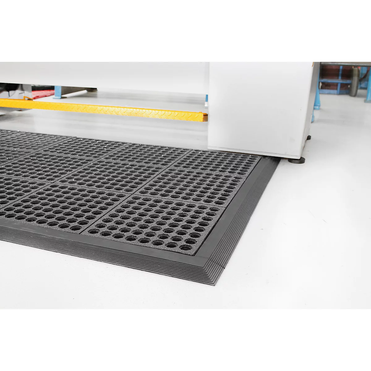 Black workstation mat with rounded edges and hole pattern. It lies in front of a machine on a white floor.