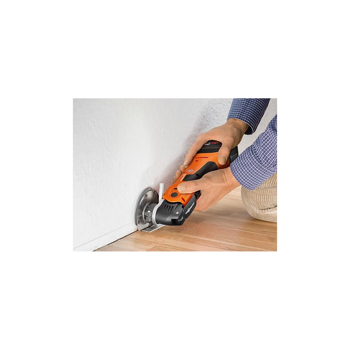 Une personne coupe dans un mur avec un multi-outil orange et noir. Du bois sur le sol.