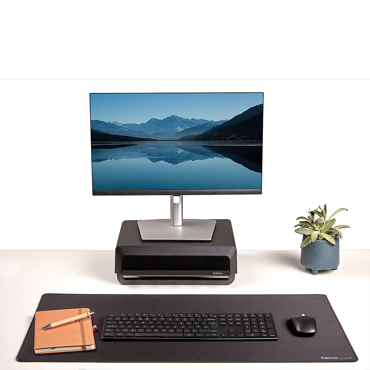 Black laptop on stand with keyboard, mouse and notebook on black mat. A small plant in the background.