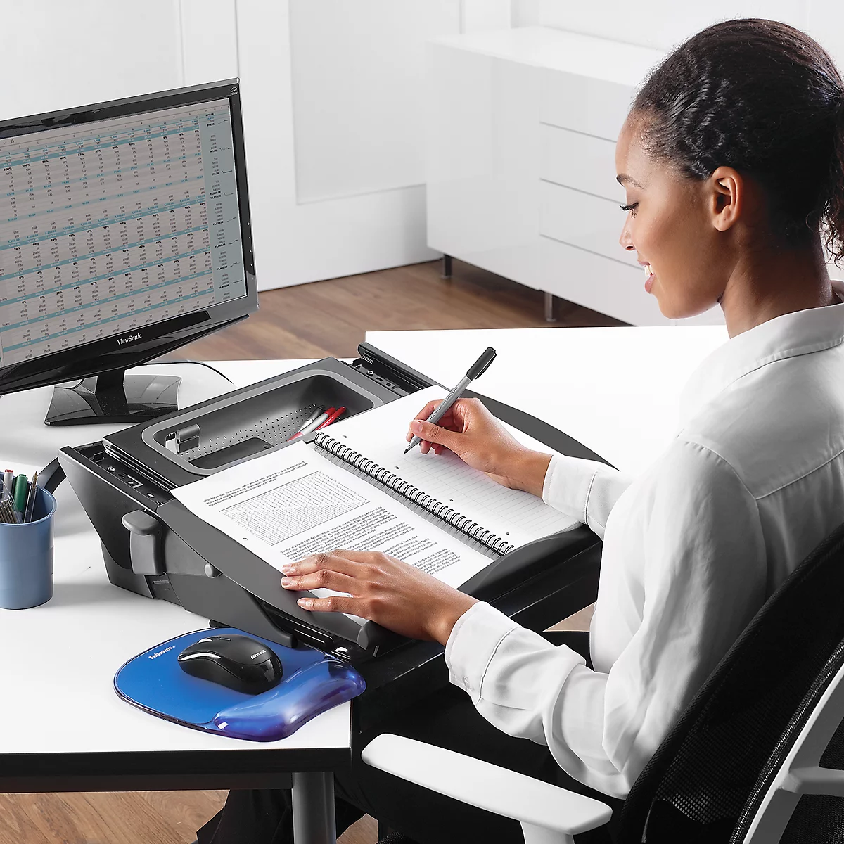 A woman writing in a notebook. She sits at a desk with a monitor and a stand that contains documents and a mouse.