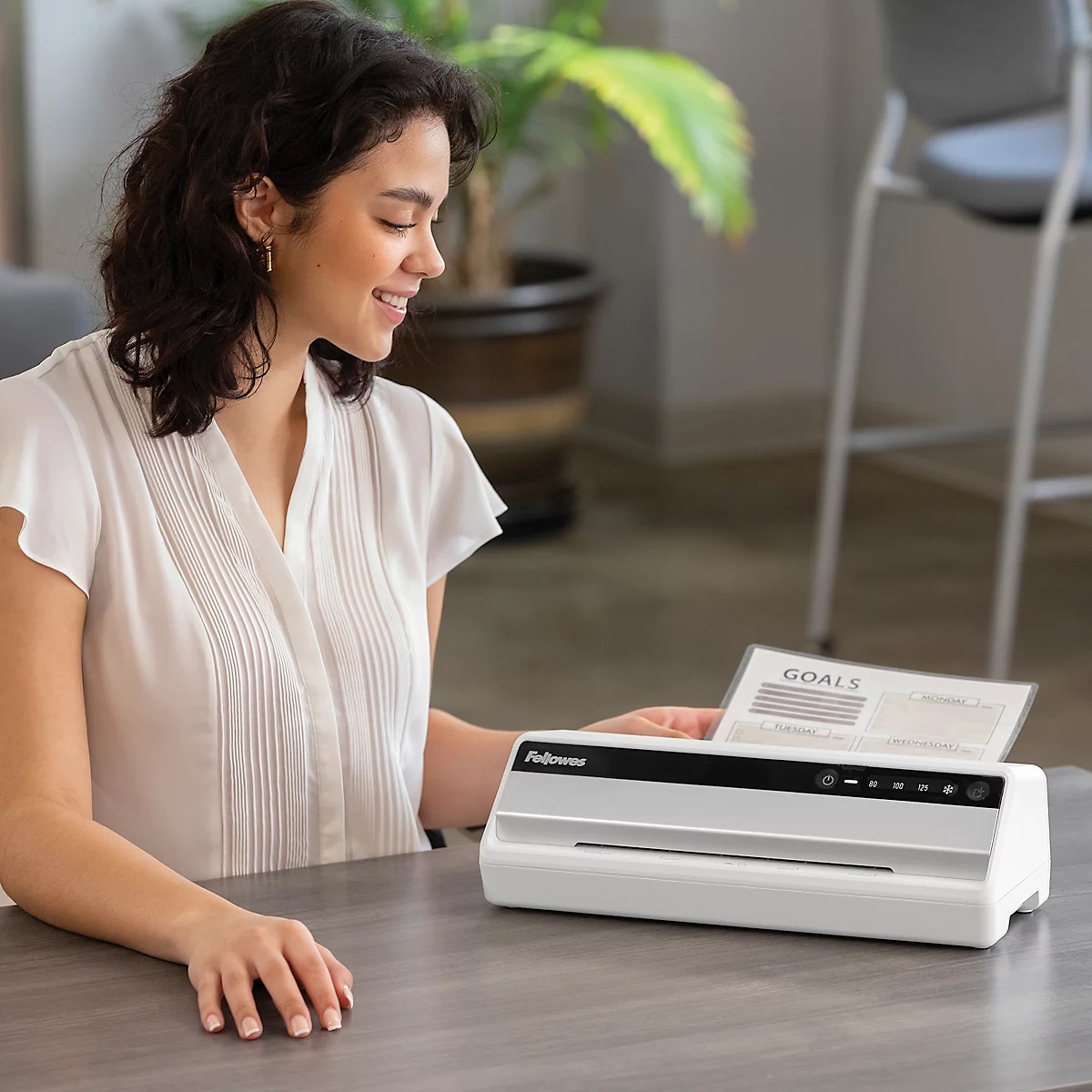 A smiling woman laminates a sheet with the word "GOALS". A white laminator is on the table.