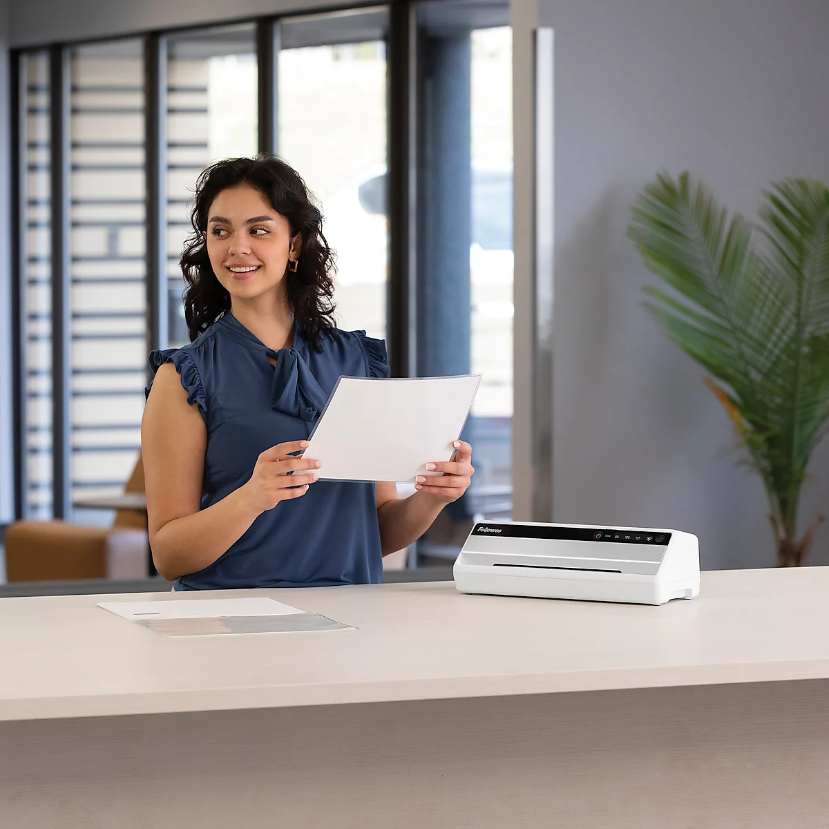 A smiling woman holds a sheet of paper at a desk. A laminator is ready.