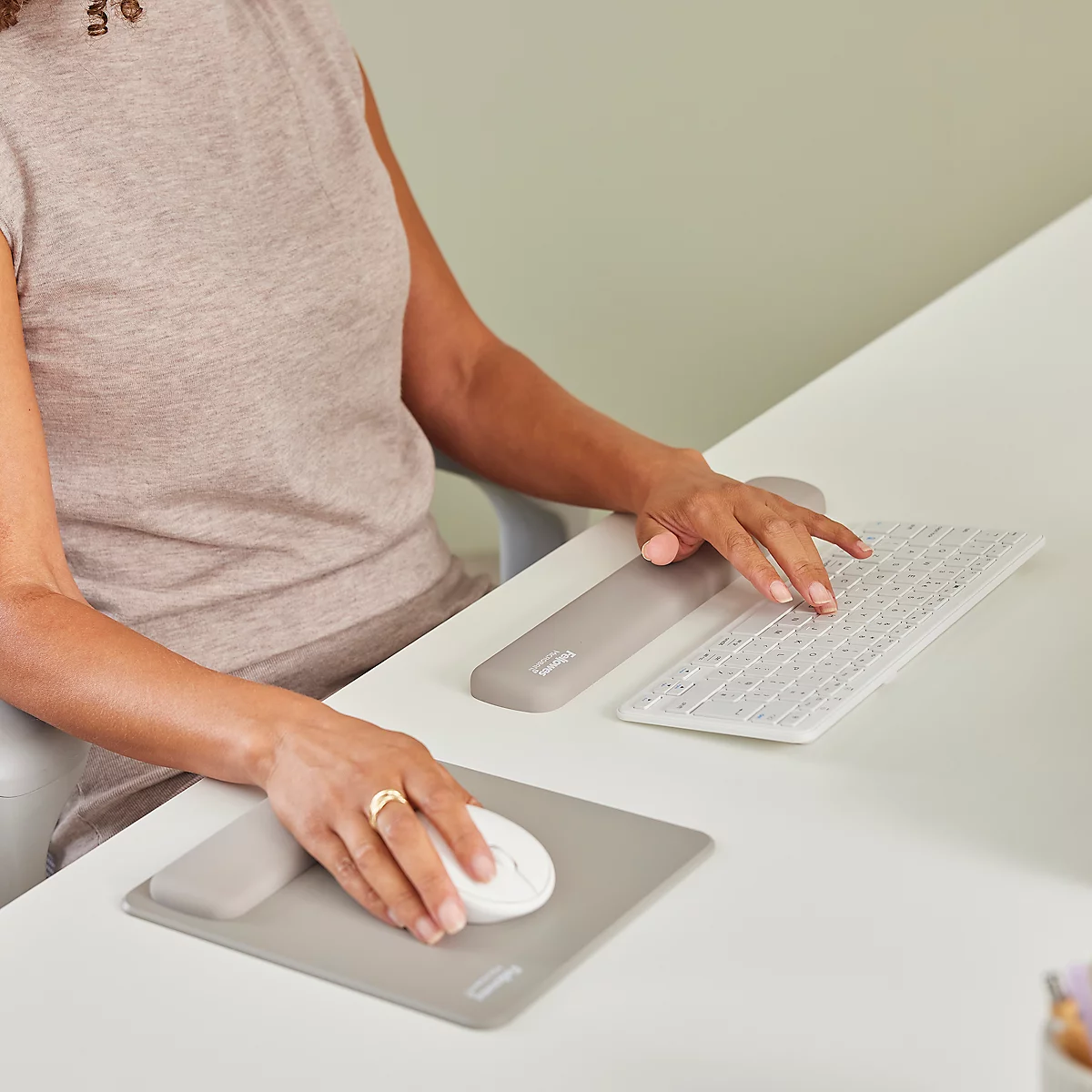 Frau sitzt lächelnd an einem weißen Schreibtisch mit Monitor. Sie trägt ein beiges Kleid und sitzt auf einem grauen Bürostuhl.
