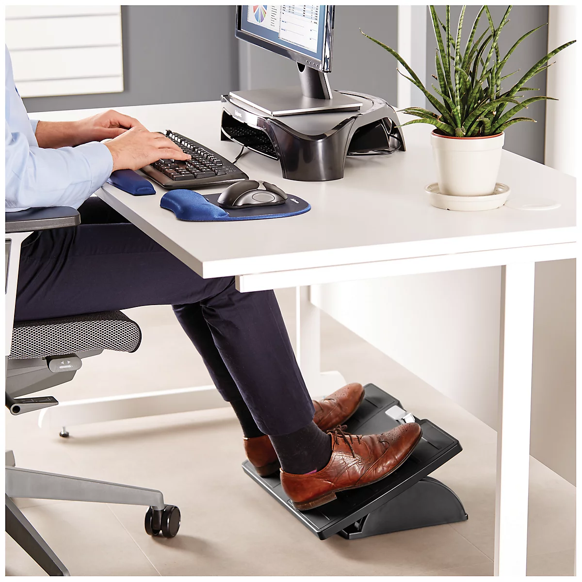 Person is sitting at the desk. Feet on footrest, hands on keyboard. Screen, plant.