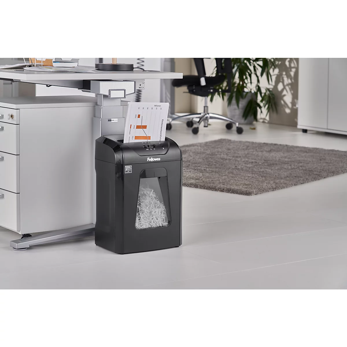 A black paper shredder stands next to a desk. Paper is being shredded at the top, and the container below is filled with shredded paper.