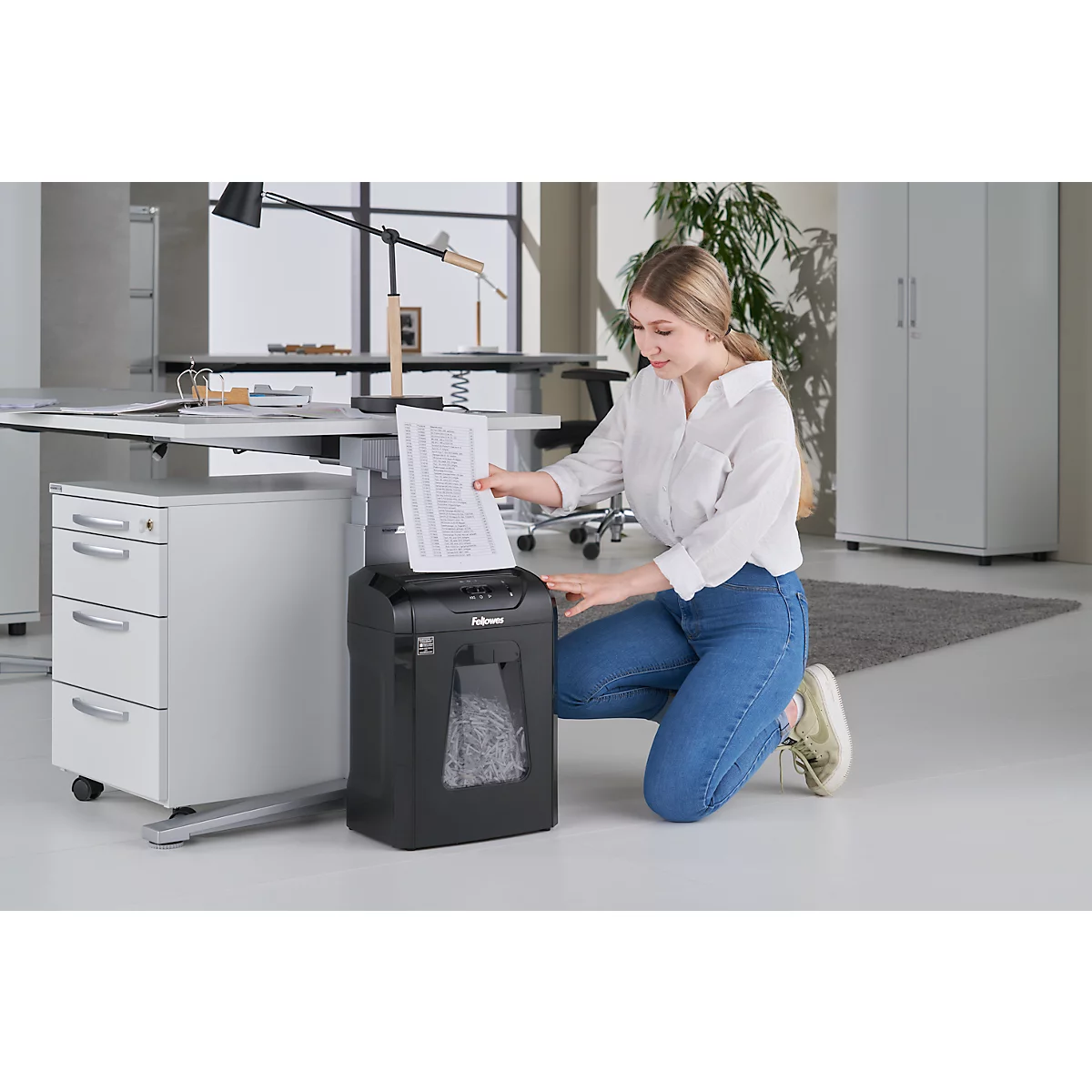 A young woman kneels, feeding paper into a shredder.