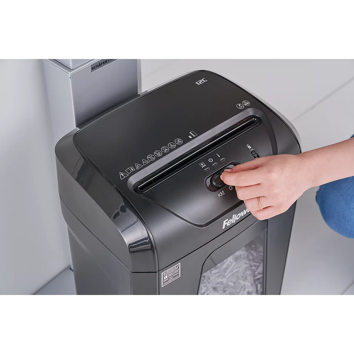 A person operates a black paper shredder. The hand is turning a knob. Shredded paper is already in the collection bin.