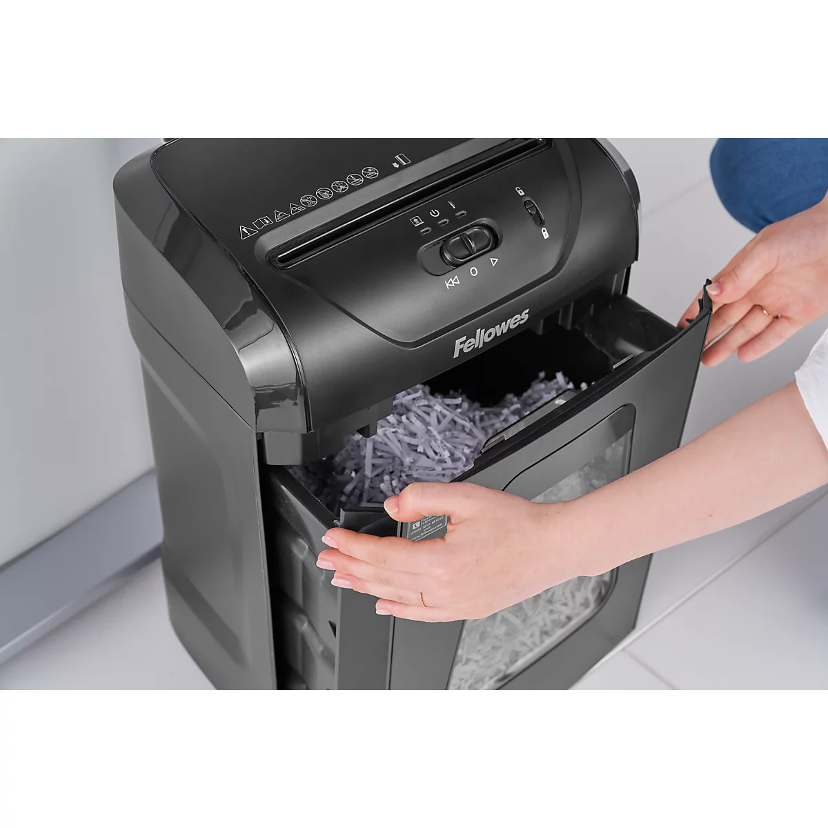 Person opening the waste bin of a black paper shredder. The bin is filled with shredded paper. "Fellowes" is visible on the device.