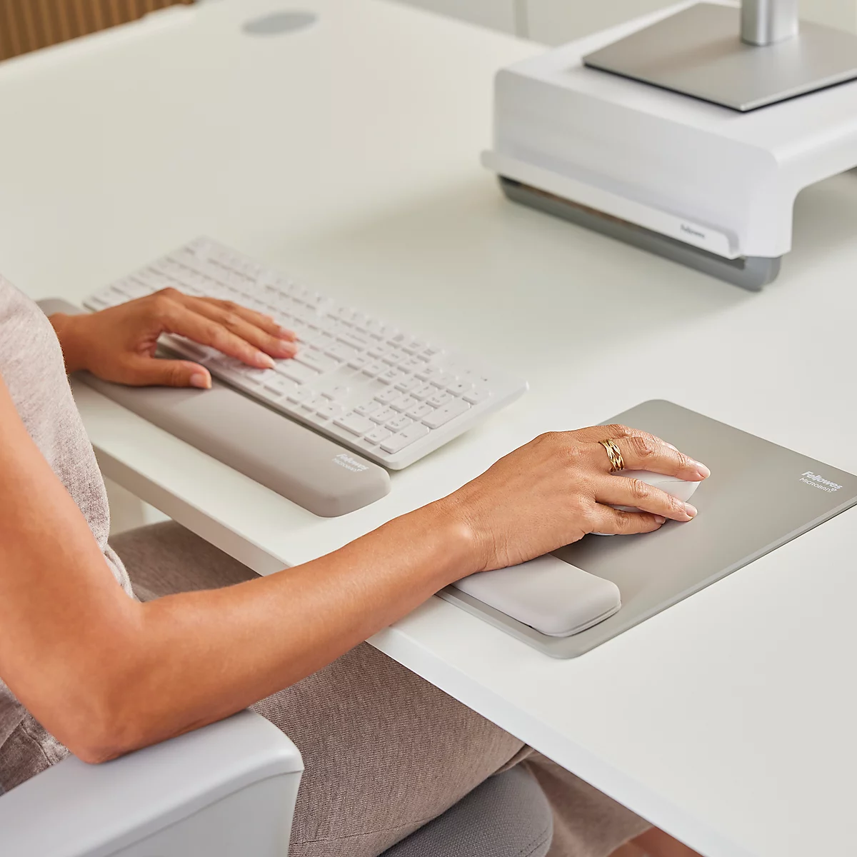 Une femme est assise à un bureau réglable en hauteur, travaillant sur un ordinateur. Elle sourit en tapant. L'espace de travail est moderne et lumineux.