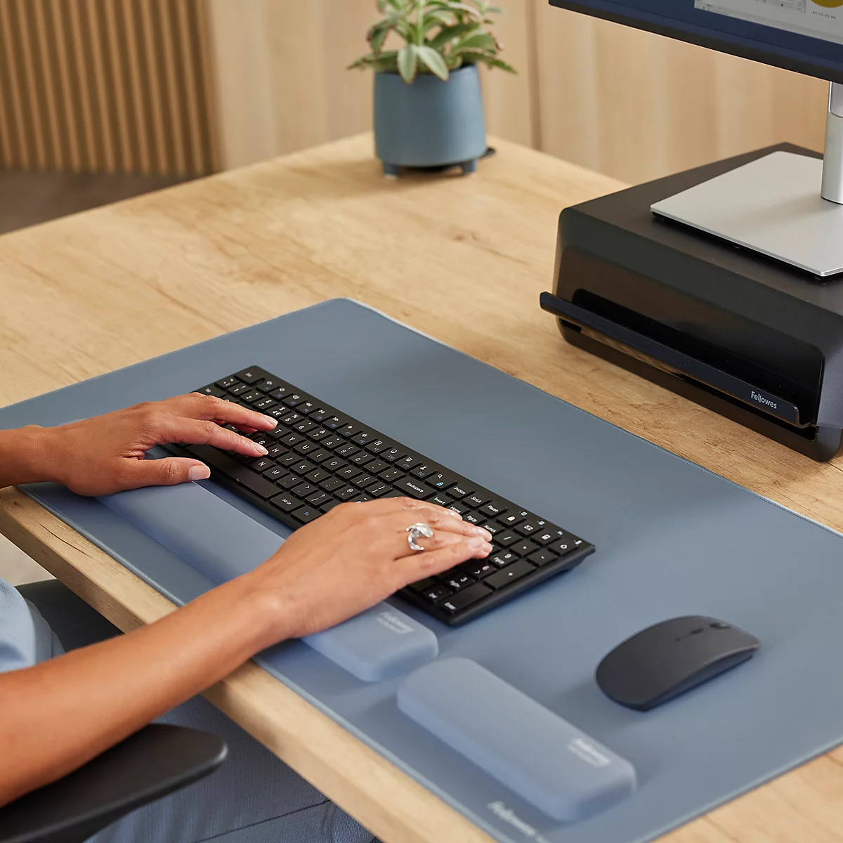 Mains tapant sur un clavier noir. Une souris et des repose-poignets sont sur un coussin bleu. Un pot avec une plante est posé sur un bureau en bois.
