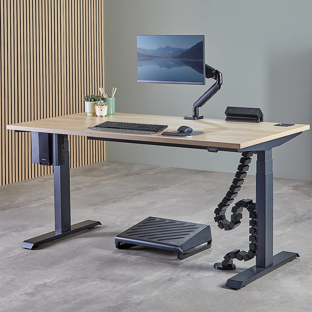 Bureau réglable en hauteur avec moniteur, clavier, souris et repose-pieds. Aspect bois, pieds gris, fond vert.
