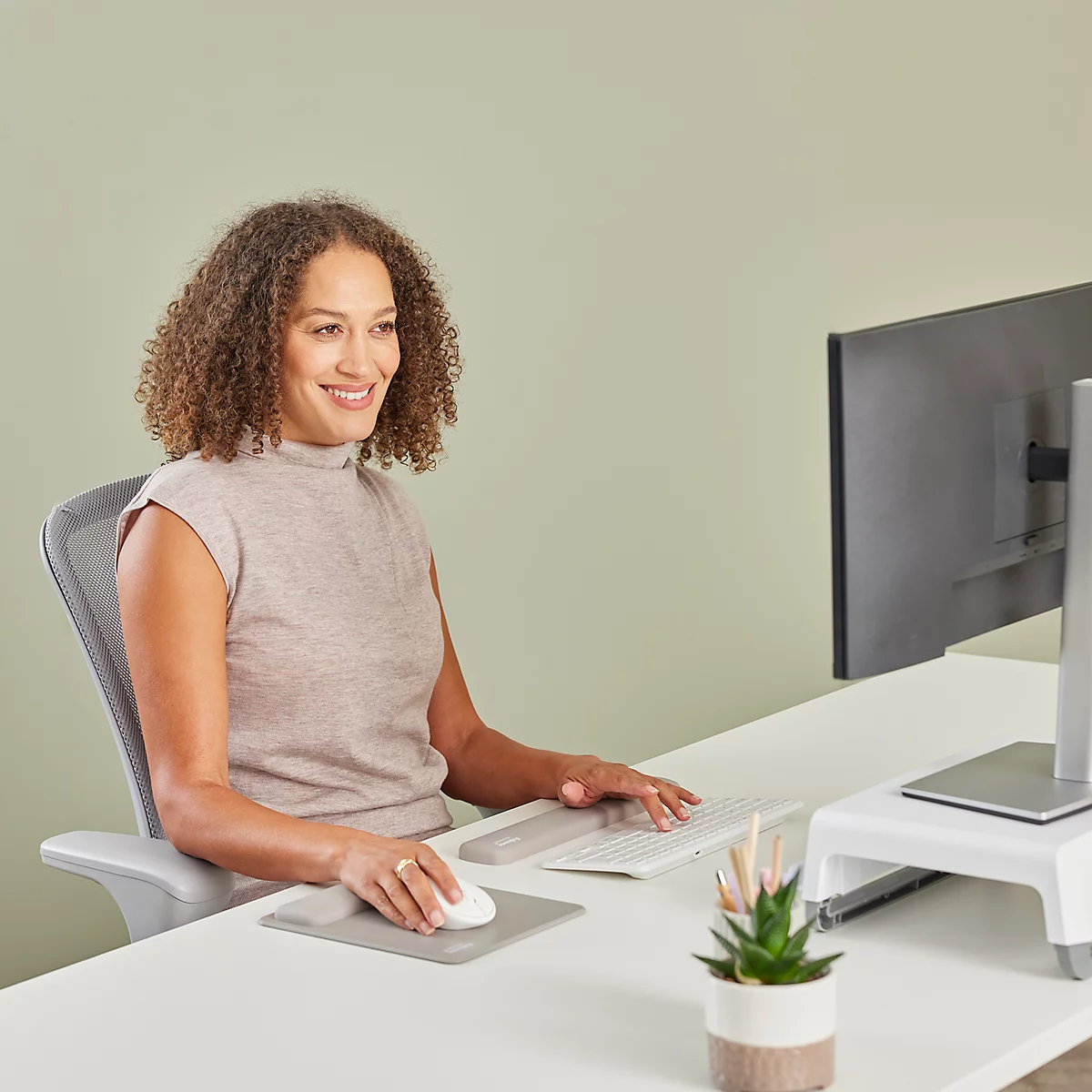 Eine lächelnde Frau sitzt an einem Schreibtisch und arbeitet am Computer. Sie nutzt eine Maus und Tastatur, während ein Monitor auf einem erhöhten Podest steht.