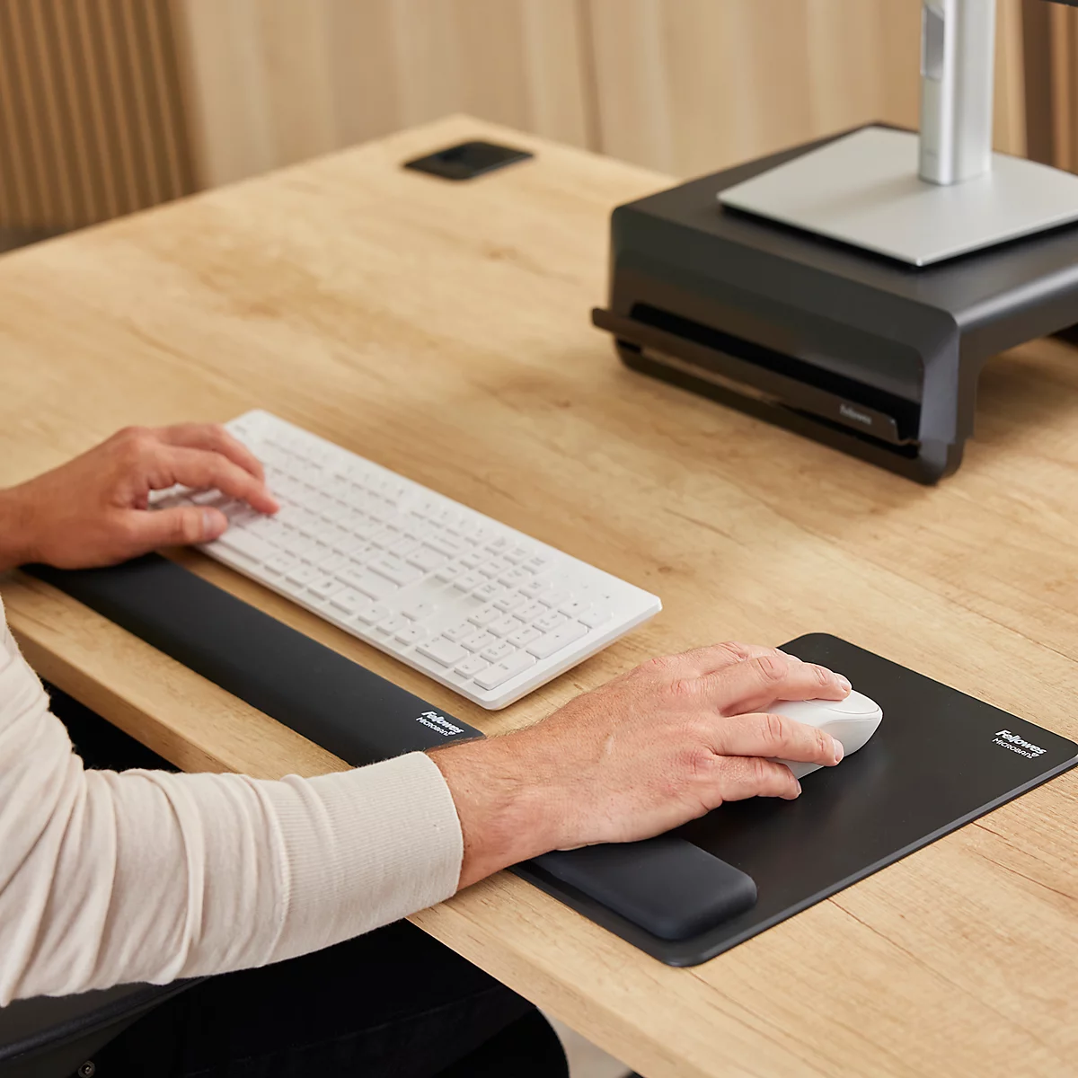 Nahaufnahme einer Person, die eine weiße Tastatur und Maus auf einem Holzschreibtisch benutzt. Ein Mauspad und eine Handgelenkauflage sind ebenfalls vorhanden.