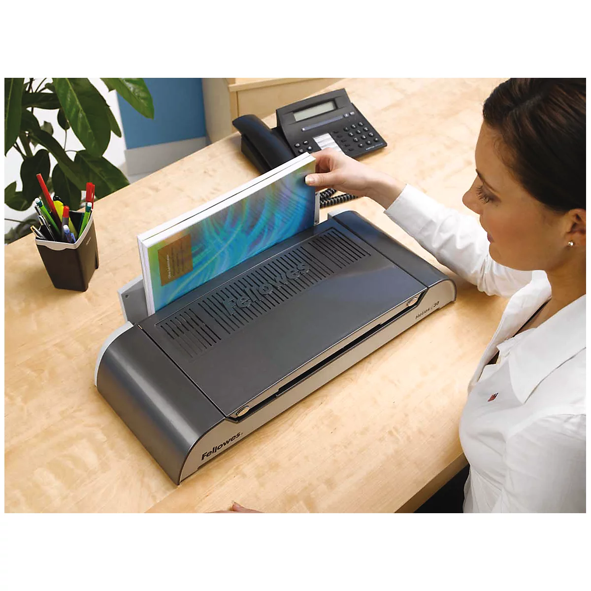 A woman laminates a document with a black laminator on a wooden desk. Pen holder on the top left, phone on the right.