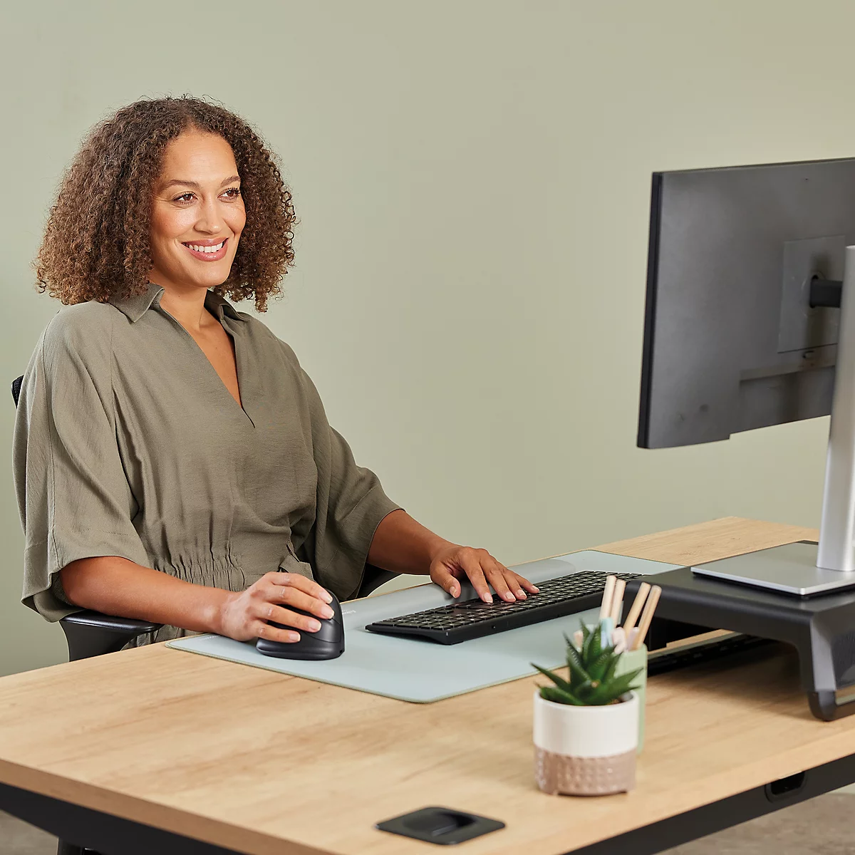 Eine Frau arbeitet an einem Schreibtisch mit Computer und Lächeln. Auf dem Schreibtisch: Maus, Tastatur, Pflanze und Stifte.
