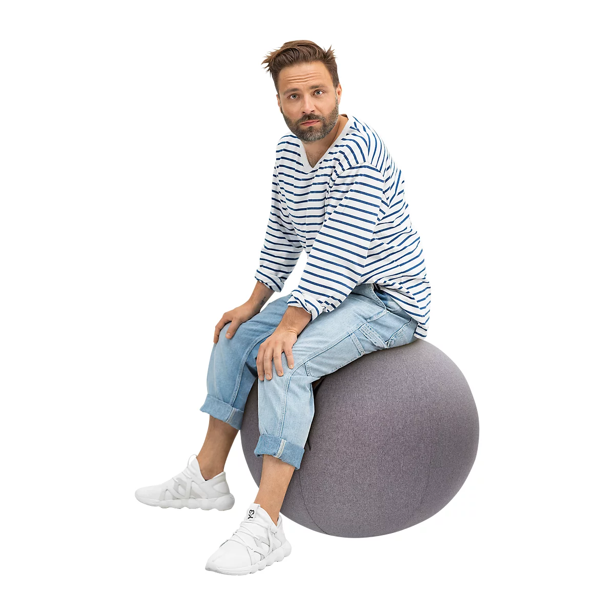 A man in a striped shirt and jeans sits on a gray exercise ball. He is looking at the camera.