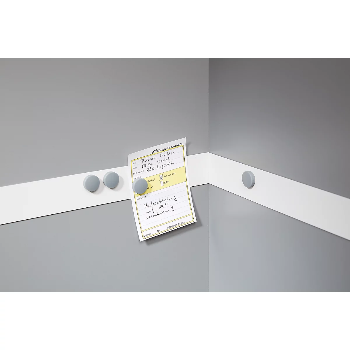Gray magnets hold a note on a white strip on a gray wall. Text on the note is visible.
