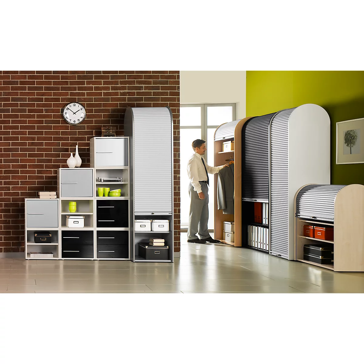 Man in office at cabinet wall, shelves to the left. Brick wall, clock, green wall.