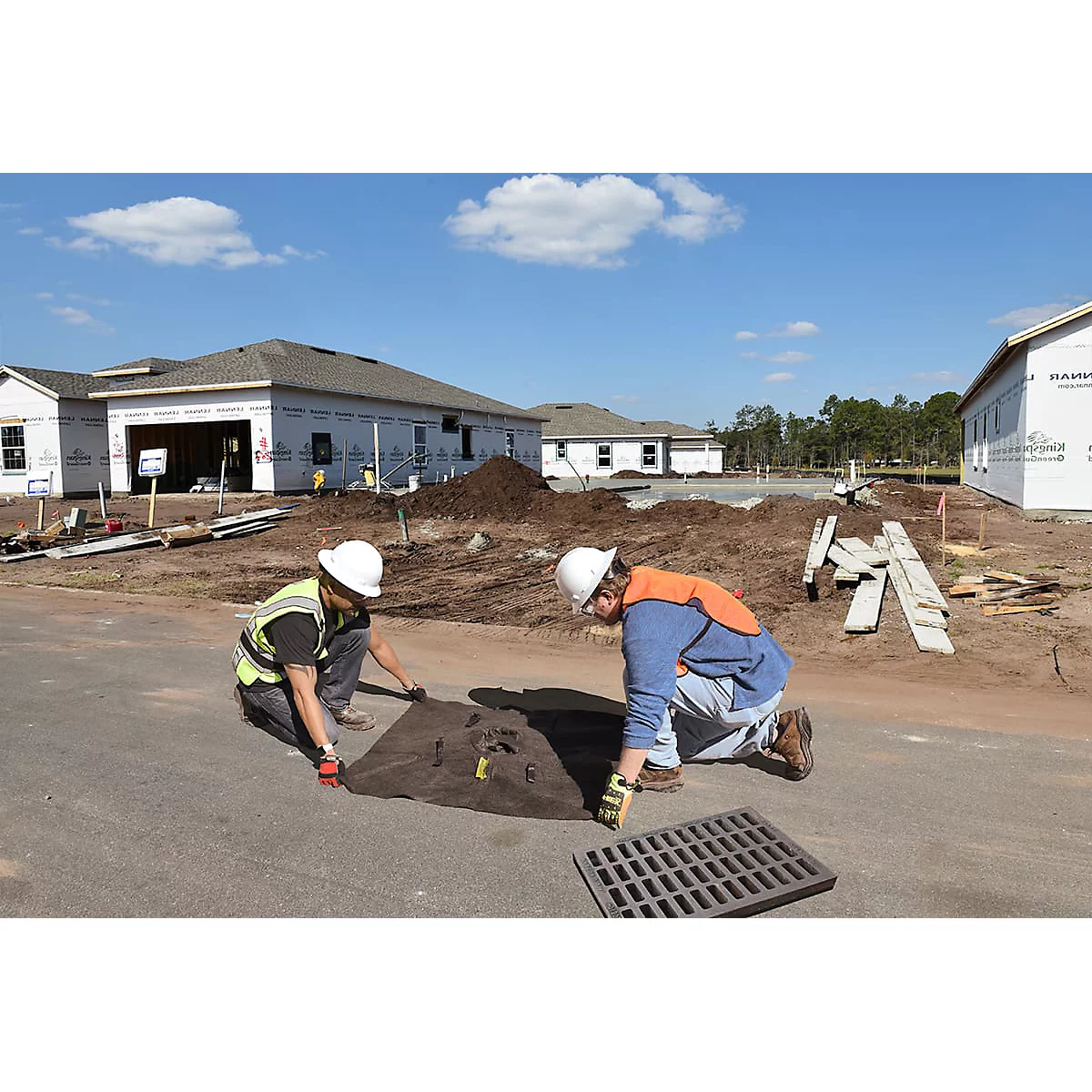 Deux ouvriers du bâtiment agenouillés sur le bord d'une route travaillant sur un drain. Des maisons en construction sont visibles en arrière-plan.