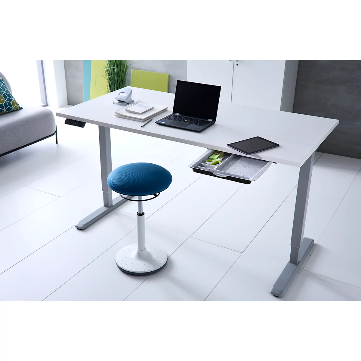 Ein höhenverstellbarer weißer Schreibtisch mit Laptop und ein blauer Hocker in einem modernen Büro.