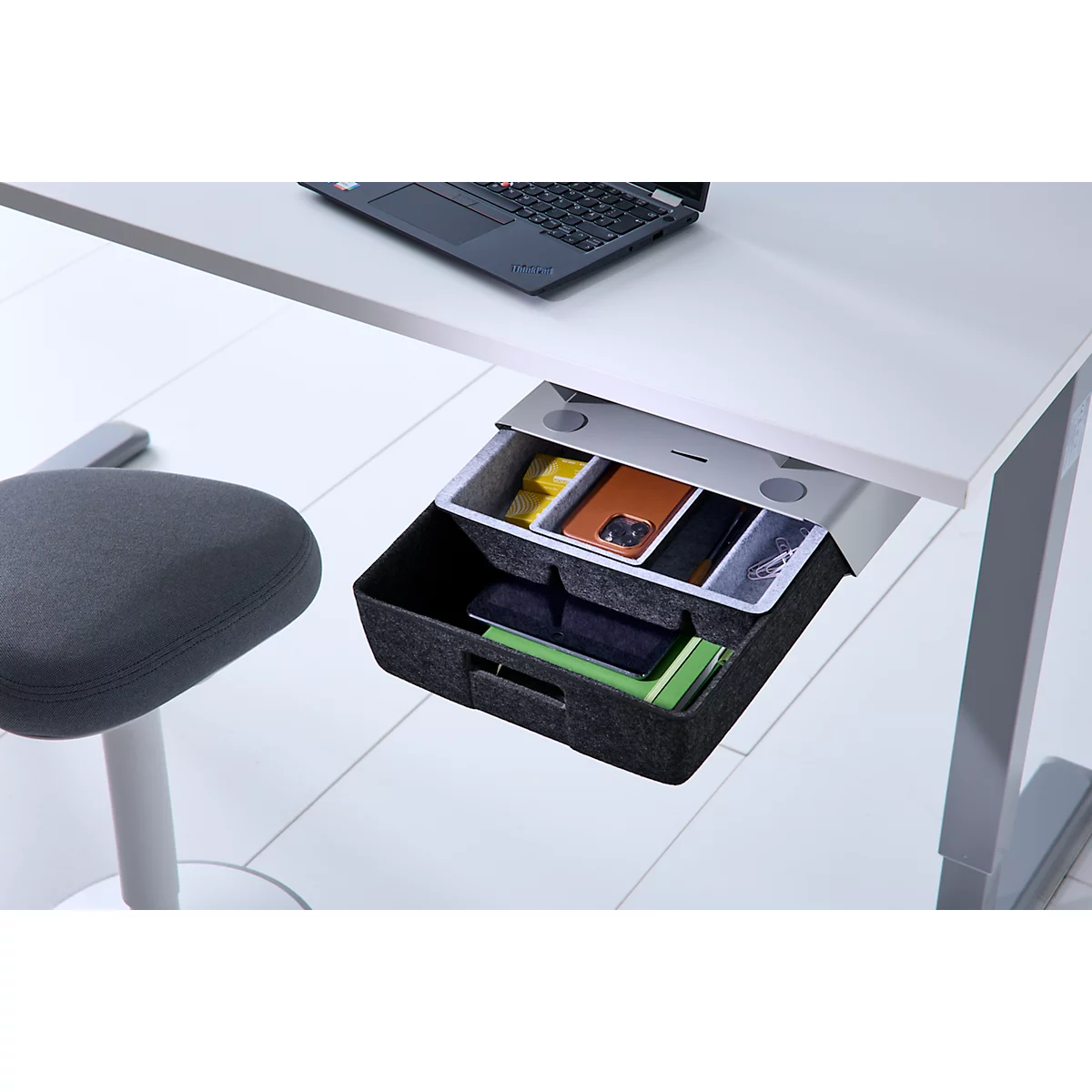 Unter dem Schreibtisch montierte, offene Schublade mit Filzeinsätzen, gefüllt mit Bürobedarf und Smartphones.
