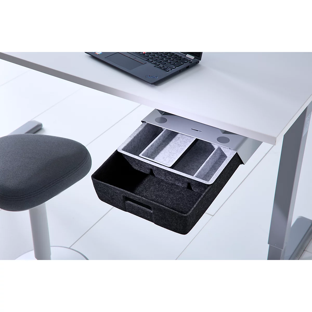 Eine offene, schwarze Filzschublade unter einem weißen Schreibtisch mit Laptop.