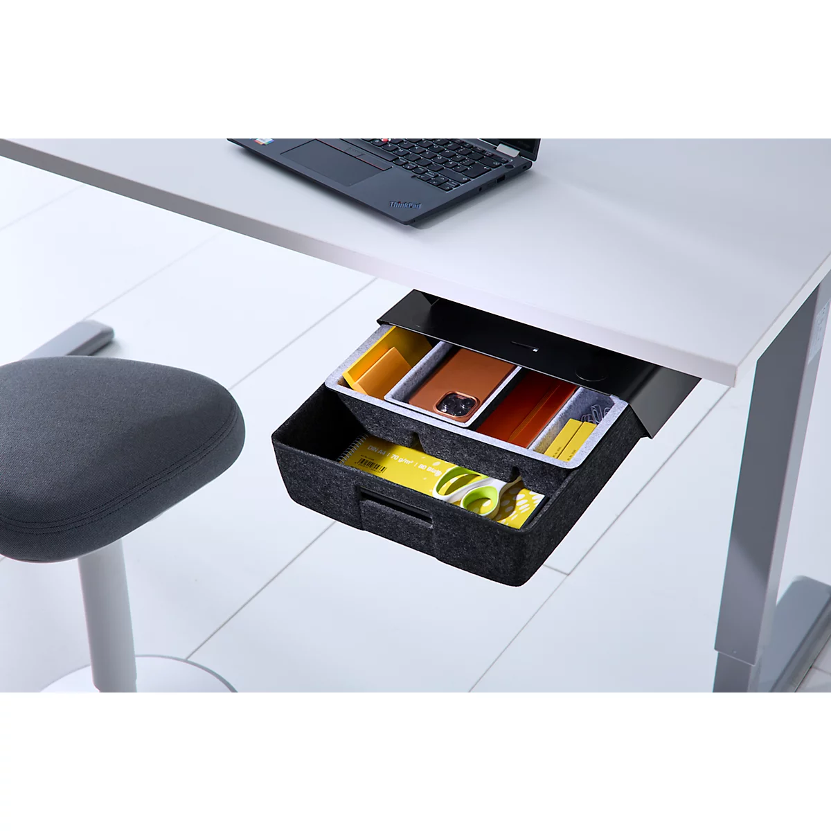 Eine geöffnete, unter den Schreibtisch montierte Filzschublade mit Büroartikeln wie Handy, Stiften und einer Schere.