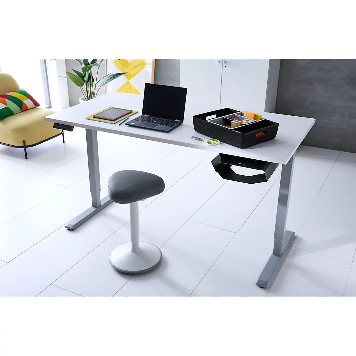 Ein moderner, höhenverstellbarer Schreibtisch mit Laptop und Ordnungsbox in einem hellen Büro.