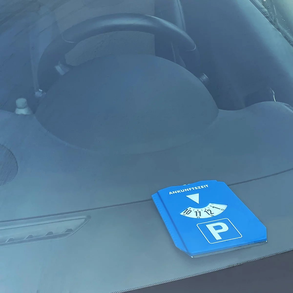 Blauer Parkausweis auf der Windschutzscheibe eines Autos. Der Ausweis zeigt die Ankunftszeit an. Im Hintergrund das Lenkrad.