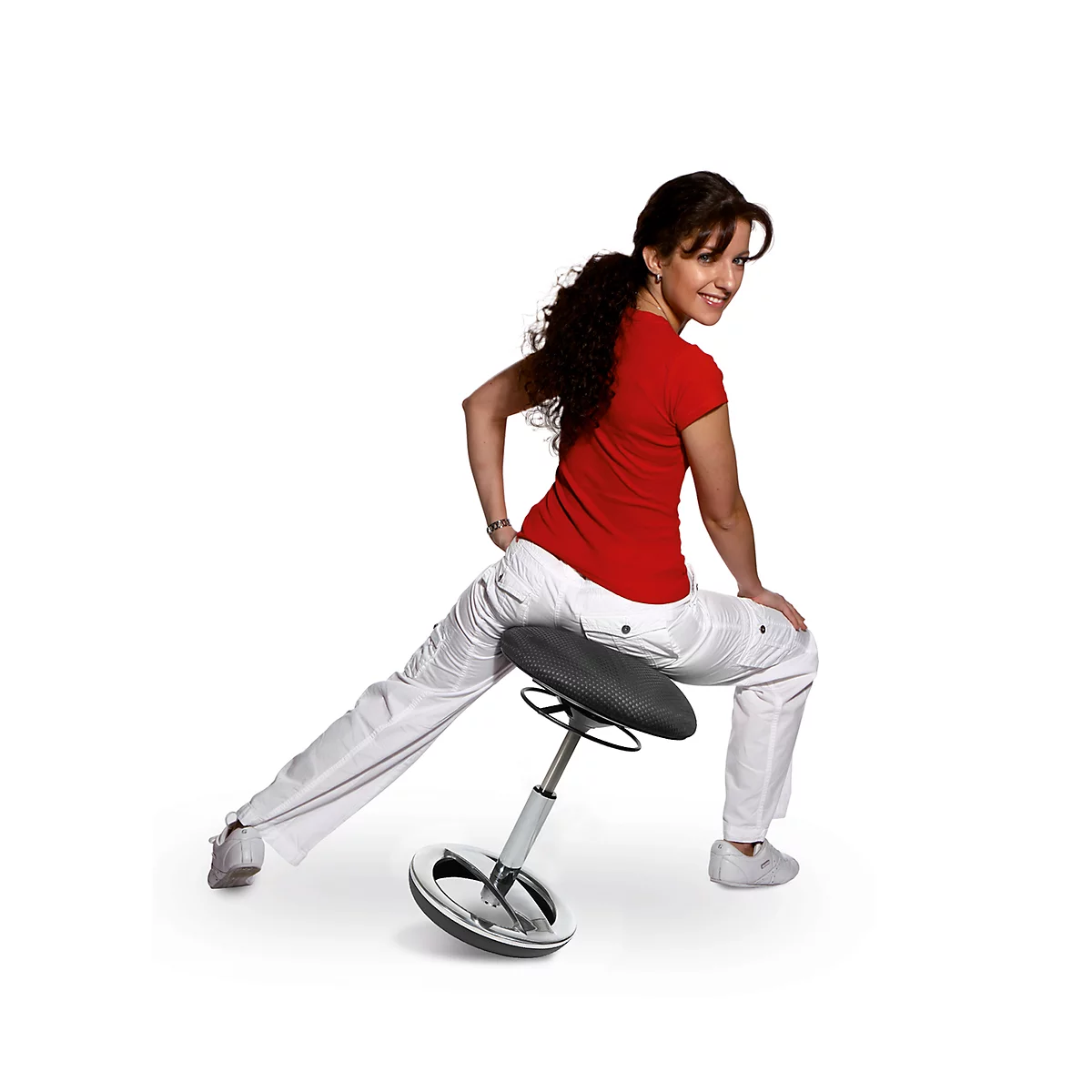 A woman in a red T-shirt and white pants sits smiling on a rocking stool.
