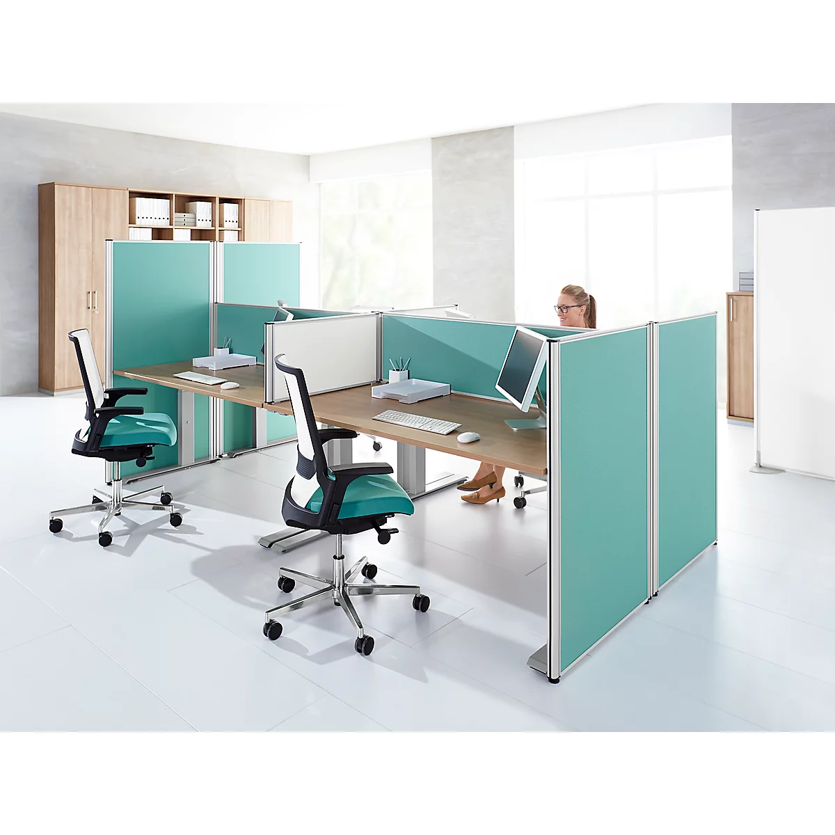 Office with desks, chairs and partitions. A person is sitting at the computer. The decor is turquoise and wood.