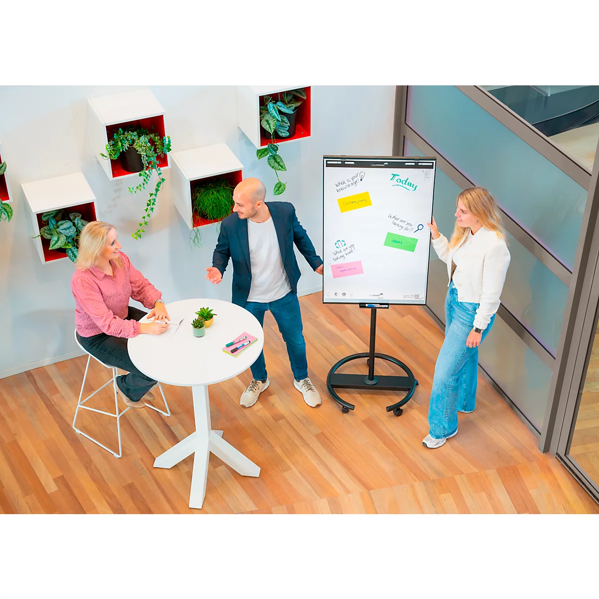 Trois personnes discutent devant un tableau blanc avec des notes. Une femme est assise à la table, deux sont debout à côté.