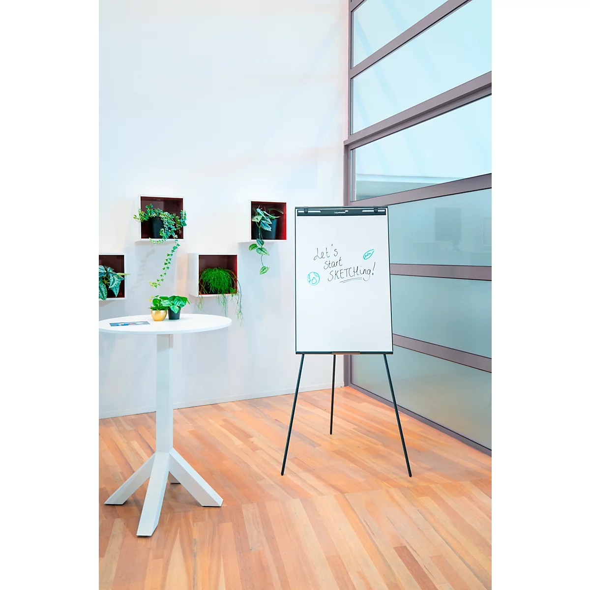 Un tableau blanc avec les mots "Let's start sketching !" est posé sur un sol en bois. Derrière, un mur blanc avec des plantes vertes.