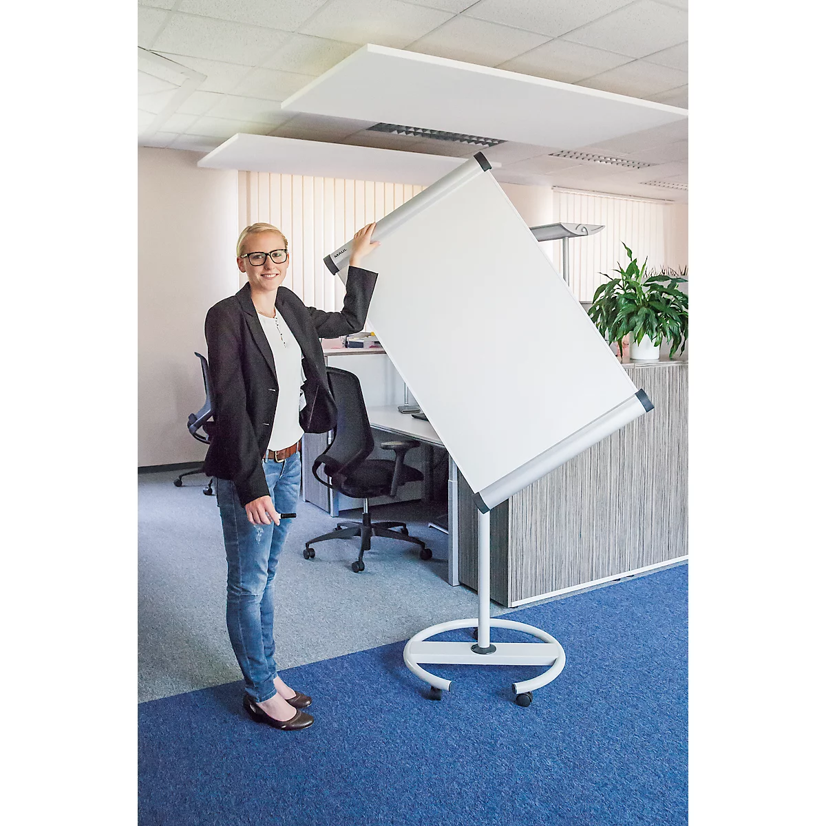 Une femme en jean et blazer tient un tableau de conférence dans un bureau. Elle sourit à l'appareil photo.