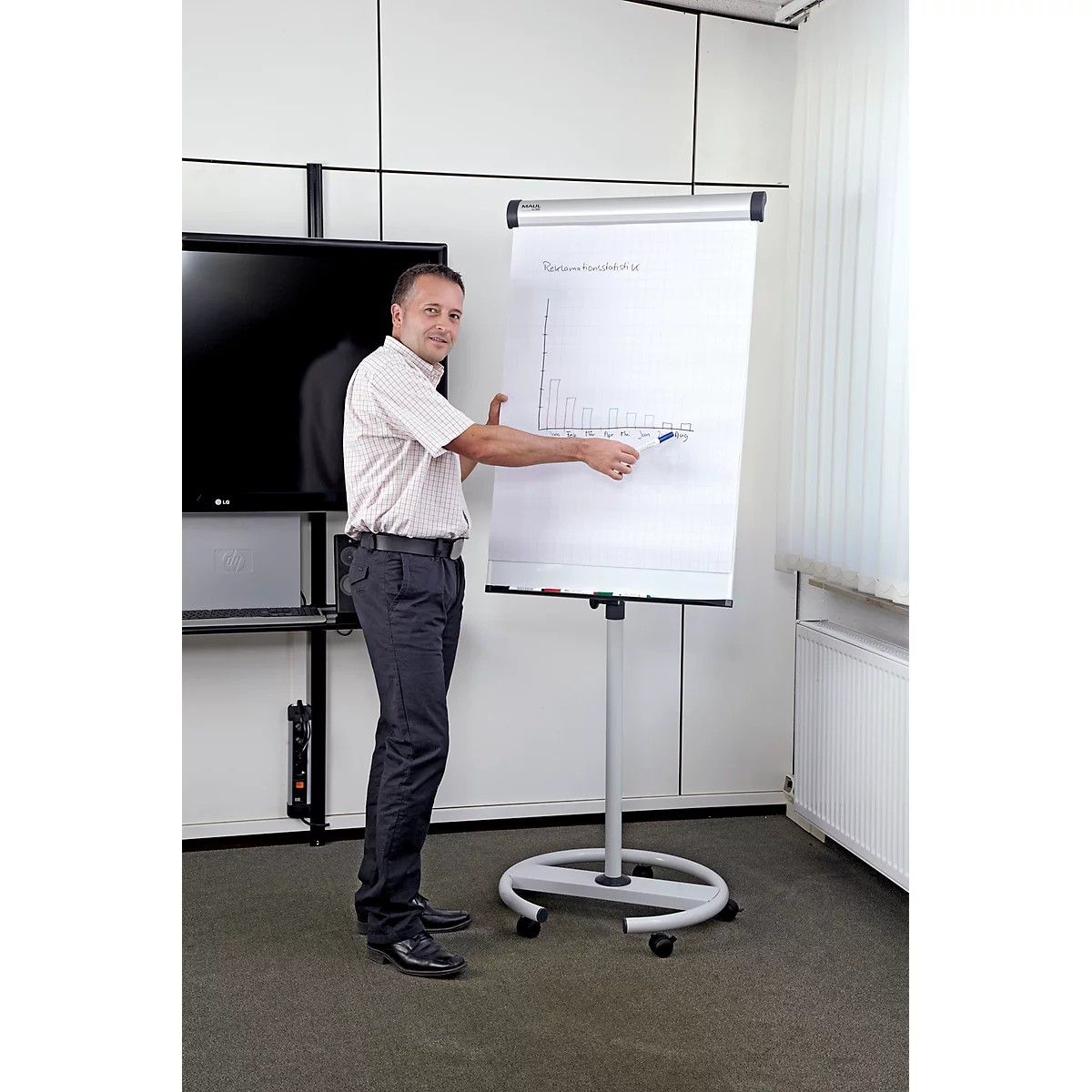 Un homme montre un graphique sur un tableau de conférence. Il porte une chemise et un pantalon. Il y a une télévision en arrière-plan.