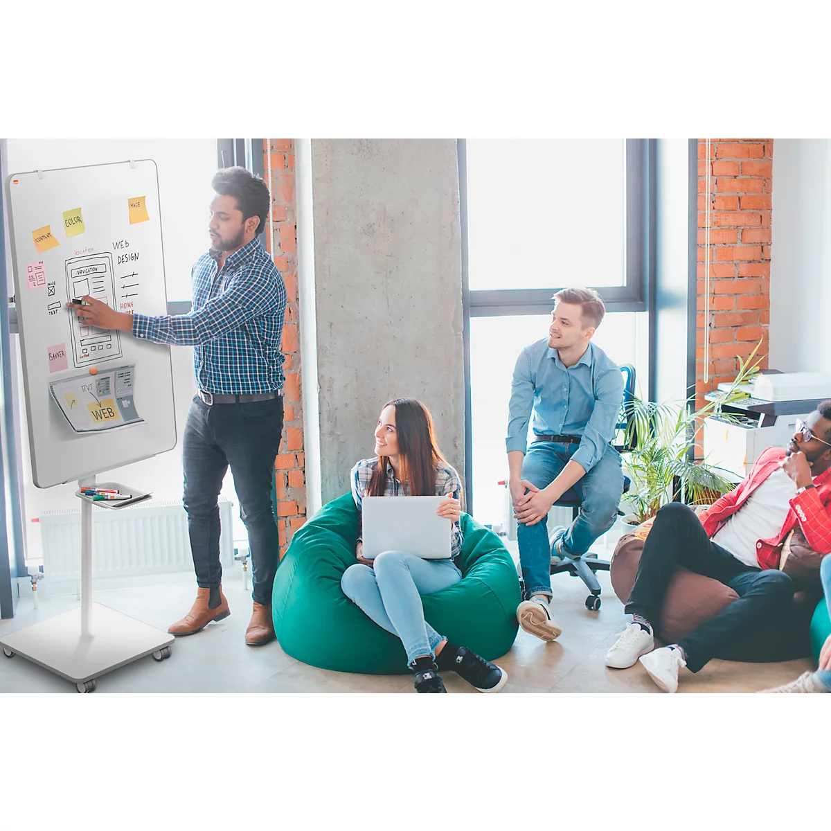 Un homme explique quelque chose devant un tableau. Une femme est assise sur un pouf vert avec un ordinateur portable.
