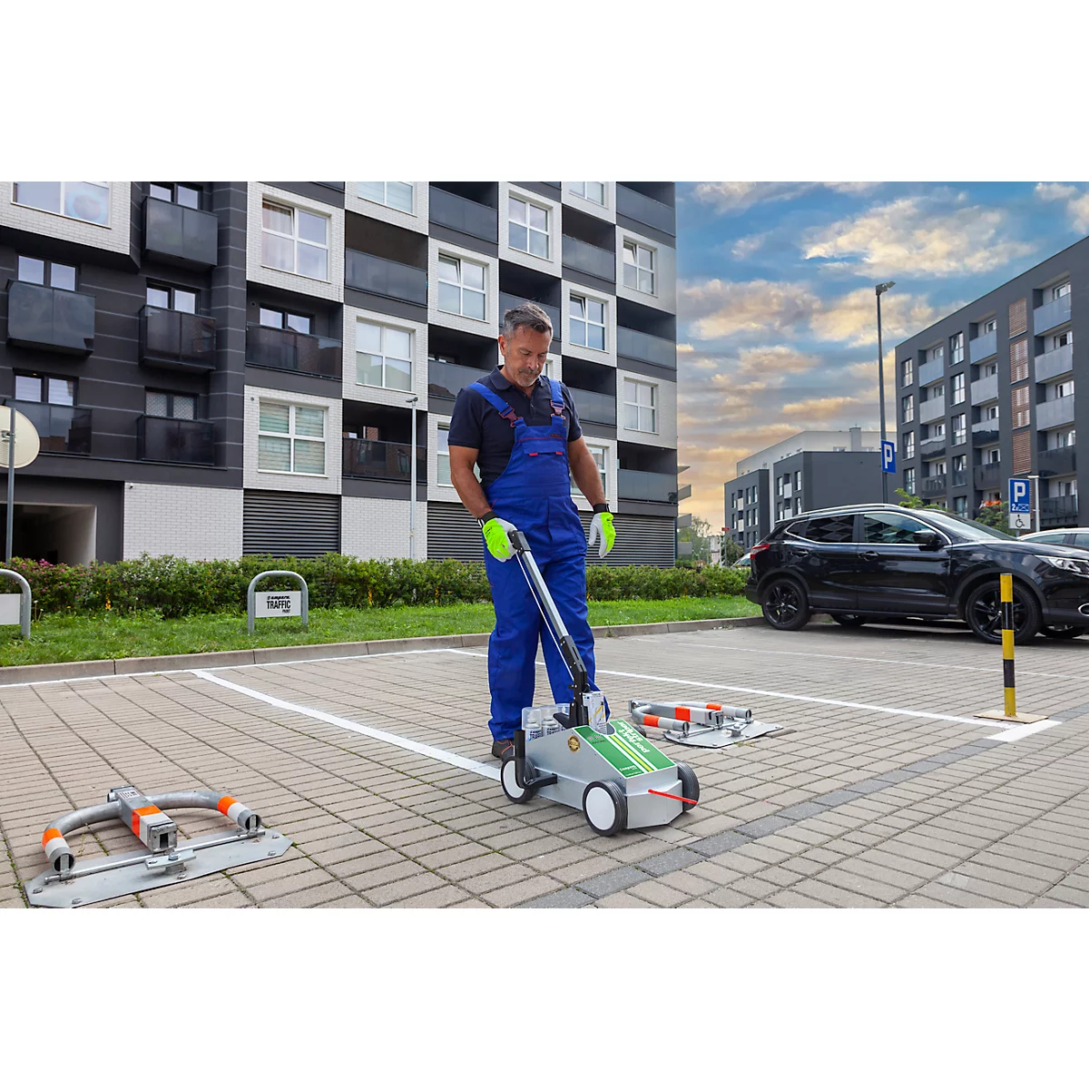 A man in work clothes operates a machine for marking parking spaces. A building and a parked car can be seen in the background.