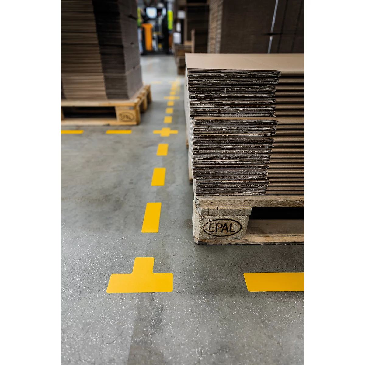 Yellow markings on a concrete floor, stacks of cardboard on pallets. Forklift and other goods in the background.