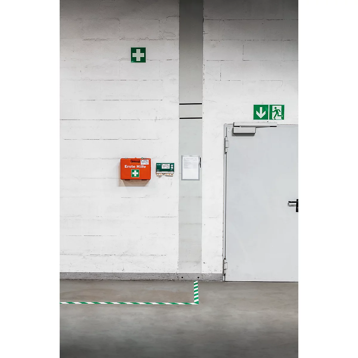 Interior shot with a white wall, featuring a first aid kit, a green cross, and an emergency exit sign. A green and white tape is on the floor.